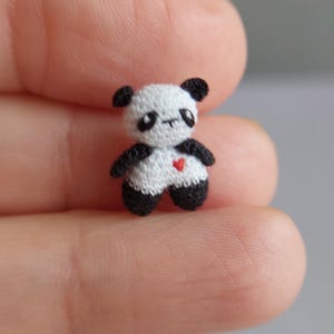 Puede incluir: Una pequeña figura de oso panda hecha a mano. El panda es principalmente blanco con detalles negros en las orejas, los brazos y las piernas. Un pequeño corazón rojo está bordado en su pecho. El panda en miniatura se sostiene entre dos dedos.