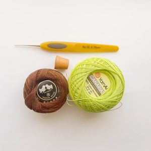 May include: A close-up of two balls of yarn, one brown and one green. The green ball of yarn is labeled "YarnA CANARIAS" and has the text "100% Cotton Mercerized 20 g / 20 г 434 m / 203 yds". A yellow crochet hook is also pictured.