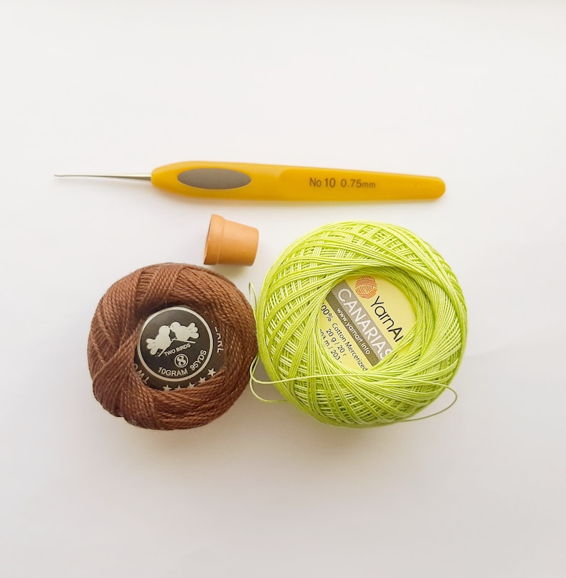 May include: A close-up of two balls of yarn, one brown and one green. The green ball of yarn is labeled "YarnA CANARIAS" and has the text "100% Cotton Mercerized 20 g / 20 г 434 m / 203 yds". A yellow crochet hook is also pictured.