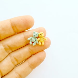 May include: Two small crocheted teddy bears, one light green and one yellow, are sitting together on a hand.