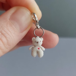 May include: A small, white crocheted teddy bear charm with a red heart on its chest. The bear is attached to a silver-colored metal clasp and ring. The charm is a miniature, handmade item.