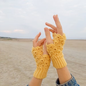 Puede incluir: Un par de guantes sin dedos de color amarillo hechos a crochet con un patrón de encaje. Los guantes se sostienen en el aire con los dedos extendidos.