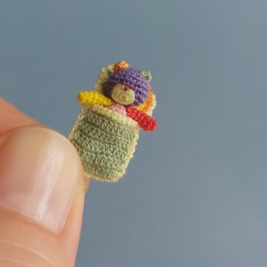 May include: A miniature crocheted teddy bear in a green blanket. The bear has a purple head, yellow arms, and a red body. The blanket is a light green color with a yellow border. The bear is approximately 0.75 inches tall.