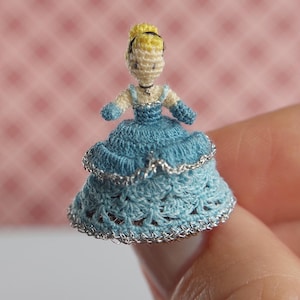 Puede incluir: Una pequeña muñeca de ganchillo que lleva un vestido azul con ribetes plateados. La muñeca tiene el pelo rubio y la cara blanca.