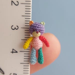 May include: A tiny crocheted teddy bear with a purple head, pink body, yellow arms, blue legs, and green feet. The bear is about 1 inch tall and is being held by a finger.
