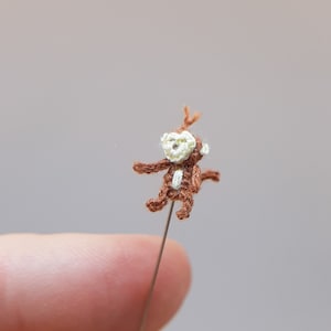 Może przedstawiać: Mały szydełkowy małpka z brązowym futrem i białą twarzą jest przypięty do igły. Małpka siedzi prosto z wyciągniętymi ramionami.