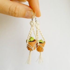 May include: A pair of white macrame earrings with small terracotta pots and green plants hanging from them.