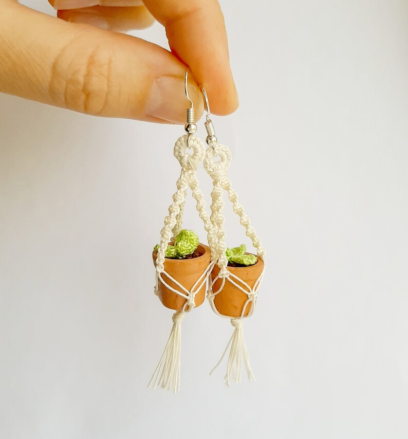 May include: A pair of white macrame earrings with small terracotta pots and green plants hanging from them.