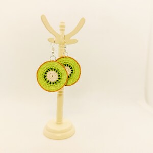 May include: A pair of crocheted kiwi fruit earrings on a white display stand. The earrings are green and brown with a white center.