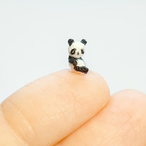 Op de afbeelding: Een kleine gehaakte pandabeer die op een vinger zit. De panda is zwart en wit met kleine zwarte ogen.