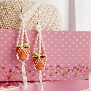 May include: A pair of handmade macrame earrings featuring small terracotta pots and green plants. The earrings are made with white string and have a tassel at the bottom.