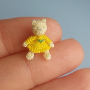 May include: A tiny crocheted teddy bear wearing a yellow dress with a green leaf design.