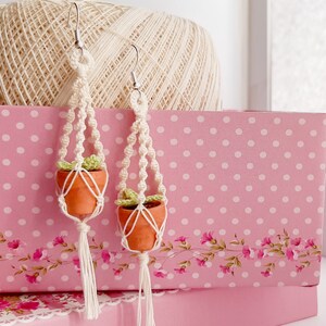 May include: A pair of handmade macrame earrings with small terracotta pots and green plants. The earrings are made with white string and have a tassel at the bottom.