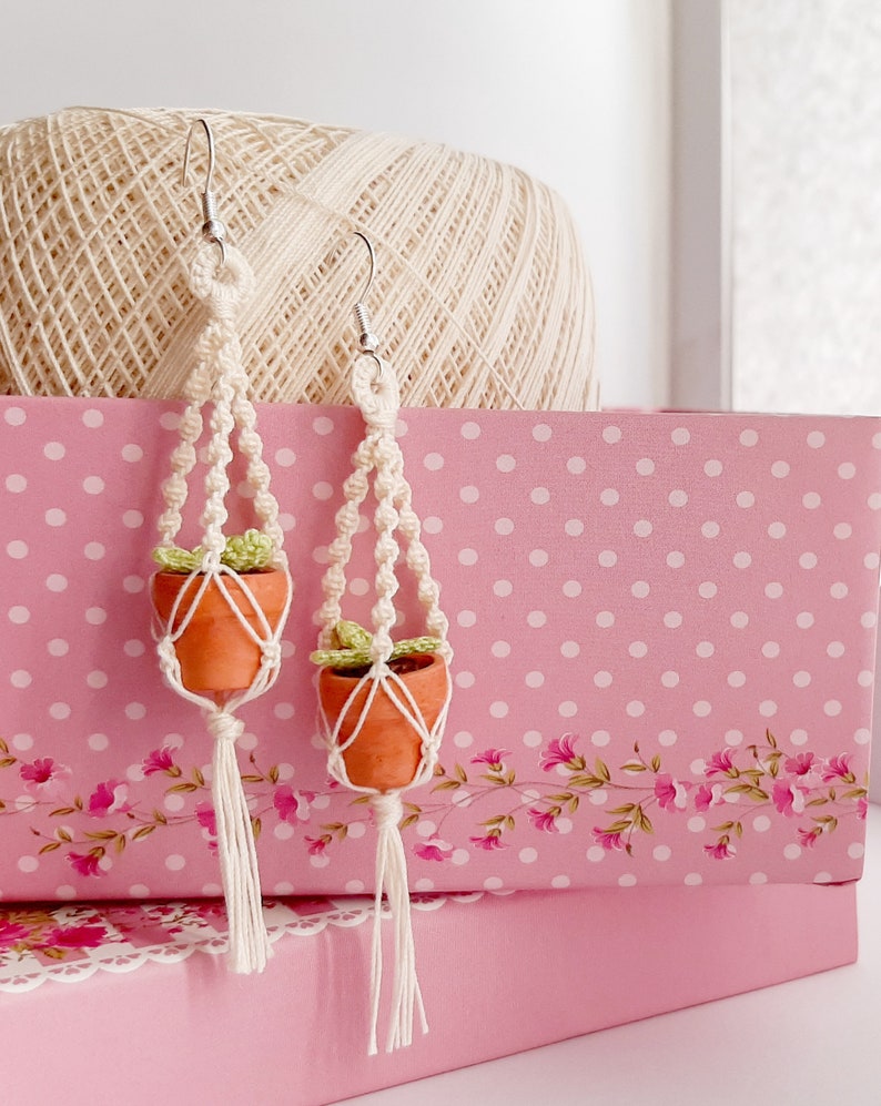 May include: A pair of handmade macrame earrings with small terracotta pots and green plants. The earrings are made with white string and have a tassel at the bottom.