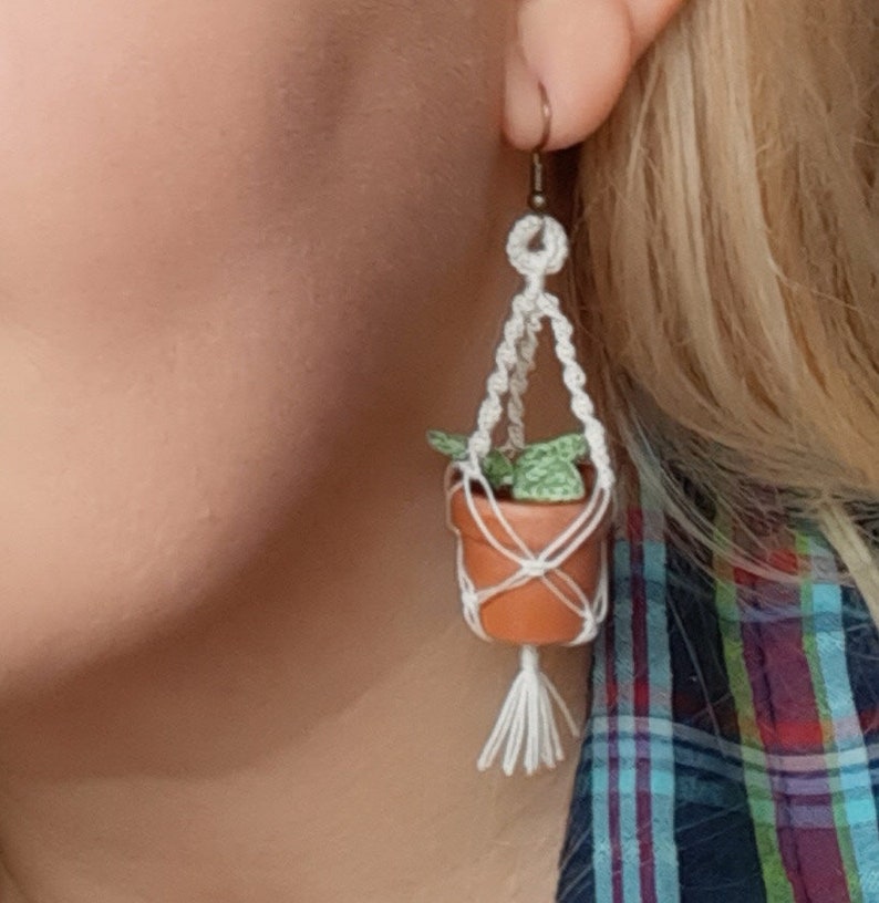 May include: A pair of white macrame earrings with a small terracotta pot and a green plant hanging from each earring.