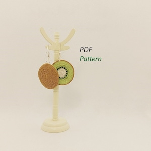 May include: Crocheted kiwi fruit earrings, one brown and one green and white, hang from a white display stand. The earrings are round with a detailed texture. The text "PDF Pattern" is visible in green.