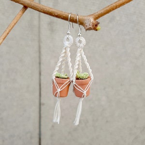 May include: A pair of white macrame earrings with small terracotta pots and green plants hanging from them.
