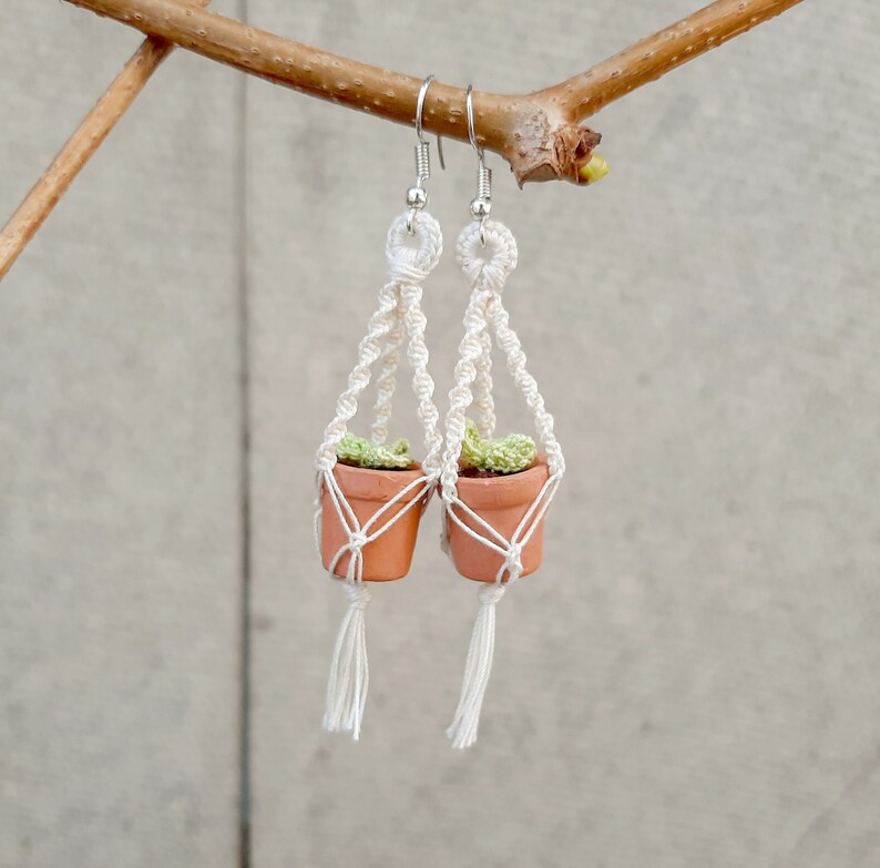 May include: A pair of white macrame earrings with small terracotta pots and green plants hanging from them.