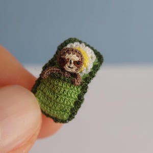 May include: A tiny crocheted sloth lying in a green blanket with a white and yellow floral detail. The sloth is brown and tan, with a smiling expression. The blanket is rectangular with a dark green edge. The background is a light blue.