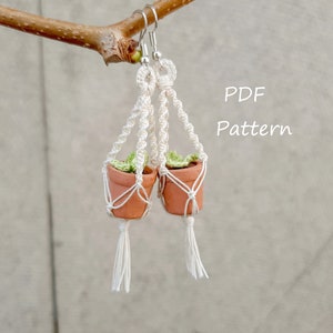 May include: A pair of white macrame plant hanger earrings with small terracotta pots and green plants. The earrings are hanging from a branch. The text "PDF Pattern" is written in white on a gray background.