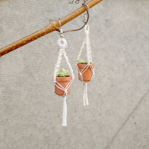 May include: A pair of white macrame earrings with small terracotta pots and green plants hanging from them.