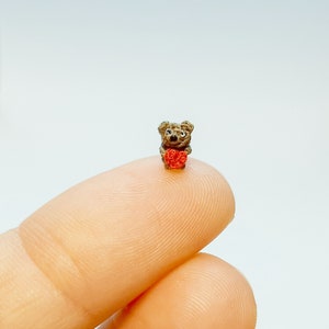 May include: A small crocheted brown bear holding a red heart. The bear is sitting on a finger.