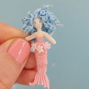以下が含まれることがあります： 青い髪とピンクの尻尾を持つ、かぎ針編みの人形。人形は花の形をしたピンクのビーズで飾られています。