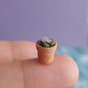 May include: A miniature terracotta flower pot with a small plant. The plant has green leaves and small purple flowers. The pot is held on a finger against a purple background. The miniature pot is a charming decorative item.