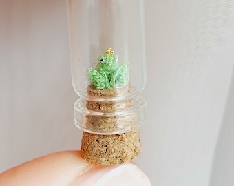 Könnte beinhalten: Eine kleine grüne gehäkelte Froschfigur mit einer goldenen Krone sitzt in einer durchsichtigen Glasflasche mit einem Korkverschluss.