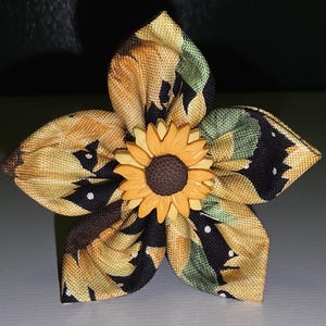 May include: A fabric flower with five petals, featuring a sunflower design. The petals are a mix of yellow, black, and green, with a small sunflower in the center. The flower is likely a decorative item.