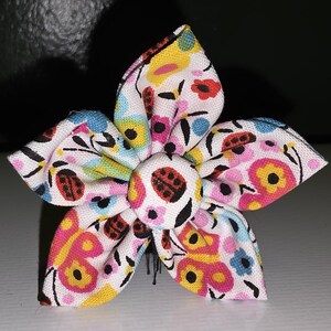 Pode incluir: Uma flor de tecido com cinco pétalas, apresentando um padrão colorido de joaninhas, borboletas e flores sobre um fundo branco. O centro da flor tem um botão com um desenho de joaninha. As pétalas são contornadas em preto.
