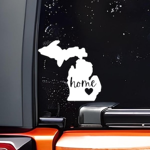 May include: White vinyl decal of the state of Michigan with the word "home" and a small heart inside the state outline. The decal is on a black car window.