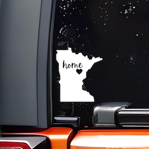 May include: White vinyl decal of the state of Minnesota with the word "home" and a small heart. The decal is on a black car window.