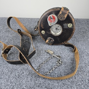 May include: A vintage black leather Detex Guardsman clock with a round face and a leather strap. The clock face is white with black numerals and hands. The clock has a metal chain and key.