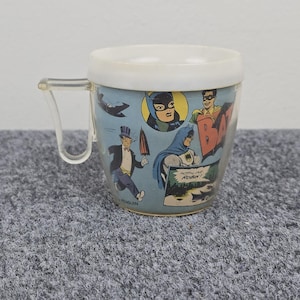 Könnte beinhalten: Vintage Batman Comic-Tasse mit transparentem Griff und weißem Rand. Die Tasse zeigt Comic-Panels mit Batman, Robin, Batgirl und dem Pinguin. Die Worte "HOT-LINE ROBIN!" sind sichtbar.