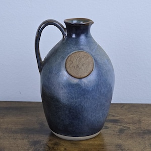 May include: A blue ceramic jug with a handle and a round, textured emblem. The jug has a dark blue rim and handle, with a lighter blue body. The emblem is a lighter brown colour. The jug is sitting on a wooden surface.