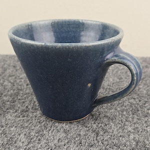 Könnte beinhalten: Handgefertigte blaue Keramiktasse mit einem einzigartigen, abgewinkelten Design. Die Tasse hat einen abgerundeten Henkel und eine strukturierte Oberfläche, ideal für Kaffee oder Tee. Die Farbe der Tasse ist ein tiefes, gedämpftes Blau.