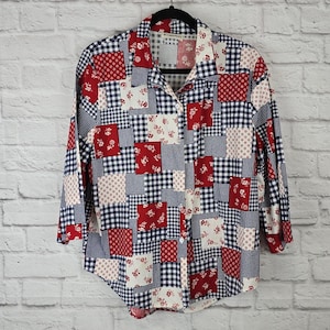 Può includere: Una camicia patchwork a maniche lunghe con colletto. La camicia presenta un mix di motivi floreali rossi, a pois bianchi e gingham blu navy. La camicia è abbottonata e appesa a una gruccia nera.