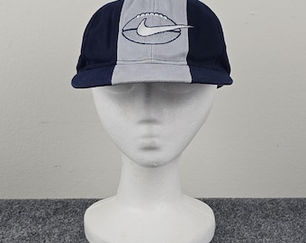 Può includere: Un cappellino da baseball blu navy e grigio chiaro con un design a forma di pallone da football e un logo Nike bianco. Il cappellino è diviso in tre sezioni verticali, con il logo Nike centrato sulla parte anteriore. Il cappellino è esposto su una testa di manichino bianca.