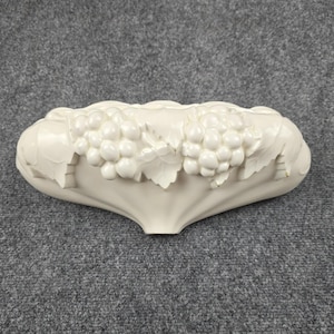 Könnte beinhalten: Cremefarbene Keramik-Wandtasche mit einem Trauben- und Blattdesign. Das ovale Dekorationsobjekt weist detaillierte Reliefarbeiten auf. Die Trauben und Blätter sind in einem realistischen Stil geformt und verleihen jedem Raum Eleganz.