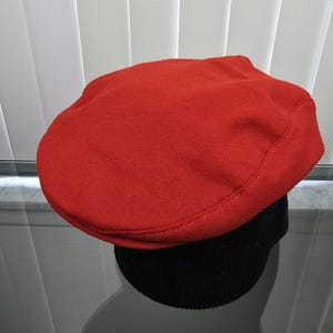 May include: A bright red flat cap with a rounded crown, sitting on a black, ribbed knit hat. The red cap is made of a soft, felt-like material. The hats are displayed on a reflective surface.