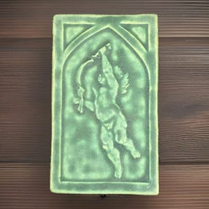 May include: A rectangular, green ceramic tile featuring a relief of a winged cherub holding a bow and arrow. The cherub is set within an arched frame. The tile has a textured surface and a light green color.