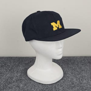 May include: A navy blue baseball cap with a flat brim. The cap features a large, gold-colored letter "M" on the front. The hat is displayed on a white mannequin head against a gray background.