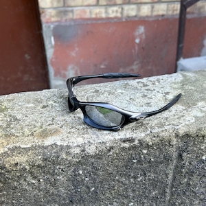 May include: Black sunglasses with a sleek, modern design. The lenses are reflective, and the frames are a matte black. The sunglasses are resting on a weathered concrete surface, with a brick wall in the background.