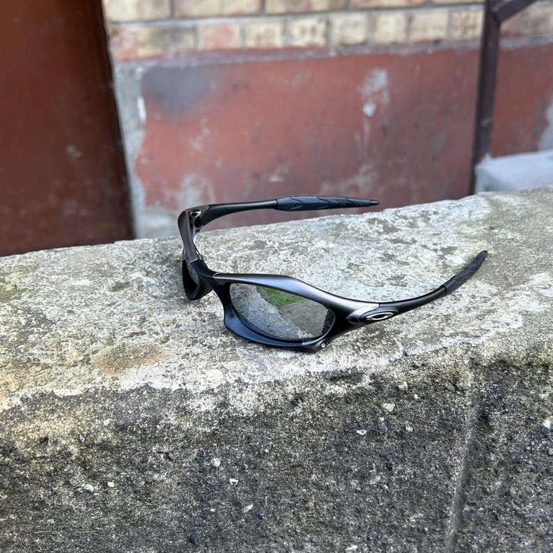 May include: Black sunglasses with a sleek, modern design. The lenses are reflective, and the frames are a matte black. The sunglasses are resting on a weathered concrete surface, with a brick wall in the background.