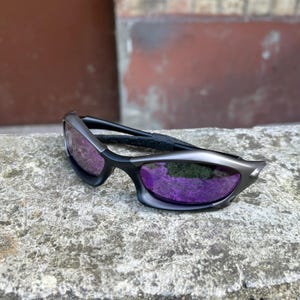 May include: Sport sunglasses with a black frame and purple lenses. The sunglasses have a sleek, modern design with a curved shape. The lenses reflect a vibrant purple color, and the frame has a matte finish.