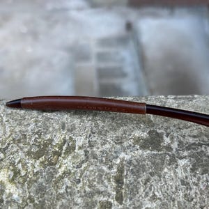 May include: Close-up of a brown eyeglass arm with a dark brown tip. The arm has a leather-like texture and the text "MADE IN U.S.A." is visible. The glasses are resting on a rough, grey stone surface.