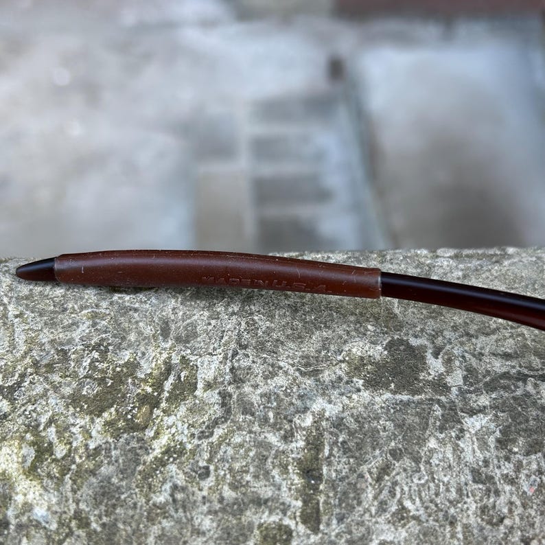 May include: Close-up of a brown eyeglass arm with a dark brown tip. The arm has a leather-like texture and the text "MADE IN U.S.A." is visible. The glasses are resting on a rough, grey stone surface.