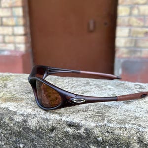 May include: Brown and black sunglasses with a curved frame and tinted lenses. The sunglasses have a sleek, sporty design with a small silver accent on the temples. The arms are a lighter brown colour.