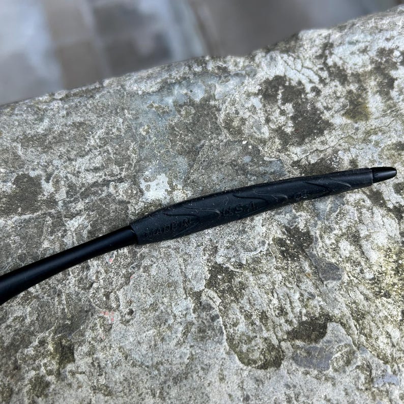 May include: A black pen with a textured grip and a pointed tip. The pen's grip has the text "MADE IN USA" embossed on it. The pen is set against a textured, gray surface.